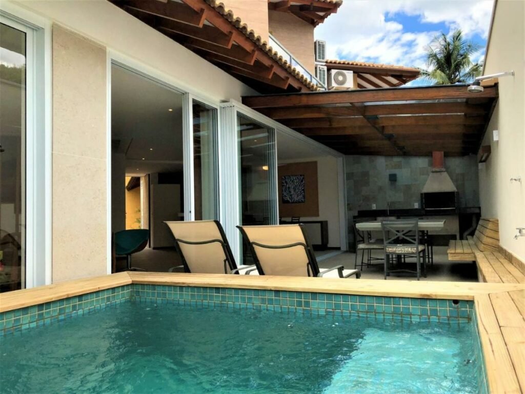 Área de lazer da Casa na praia linda de Juquehy durante o dia, com piscina a frente e ao fundo área de lazer com duas cadeiras e ao fundo uma mesa com cadeiras e ao fundo a churrasqueira.