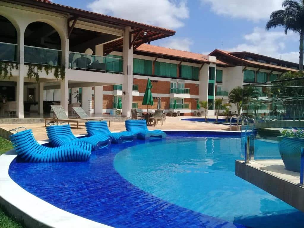 Piscina azul redonda com espreguicadeiras azuis na borda, mesas e cadeiras atrás e quartos de hotel ao fundo.
