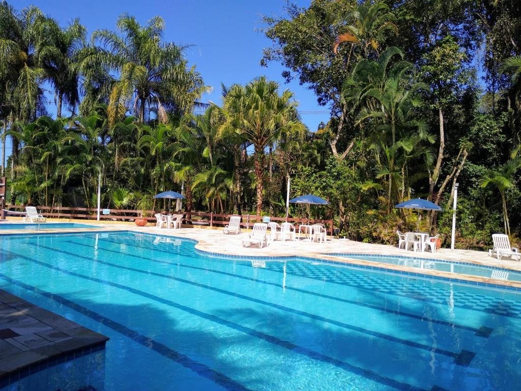 Piscina no Riviera Flat Service- 109, local amplo e cercado por árvores, coqueiros e vegetação, no deck estão algumas cadeiras, mesas e guarda-sóis