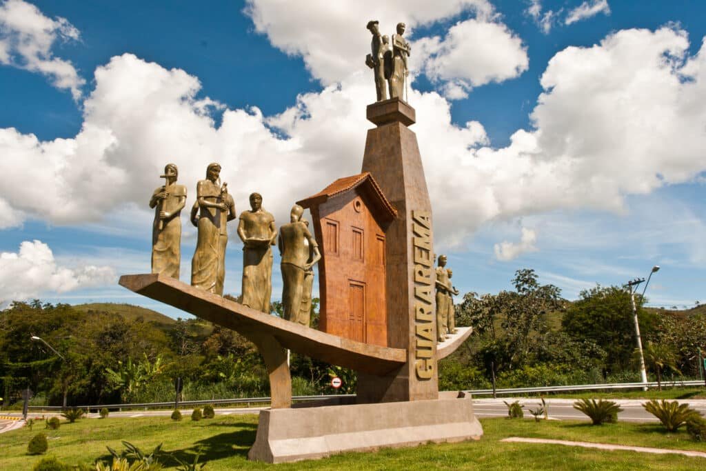 Portal origens de Guararema, com escultura de mulheres e homens e entrada de uma casa, escrito ao meio "GUARAREMA". Há um gramado baixo com pequenos arbustos em volta, uma rua asfaltada atrás e bastante natureza ao fundo