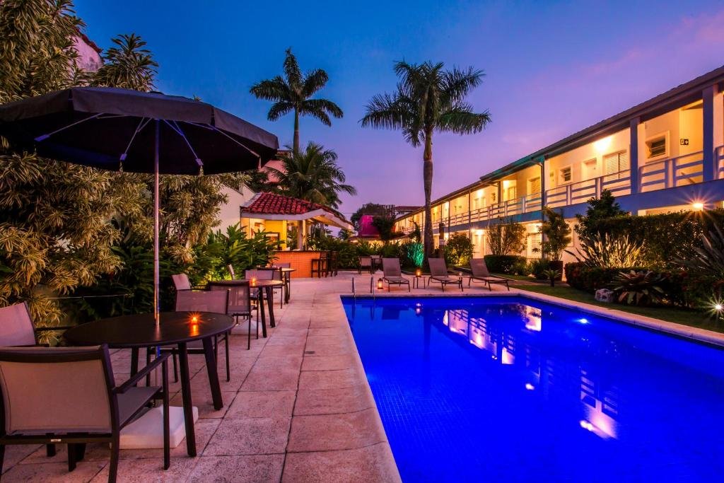 Piscina do Pousada Azul da Cor do Mar com um deck cercado por cadeiras e mesas redondas com guarda-sóis, além de algumas palmeiras e um pequeno jardim