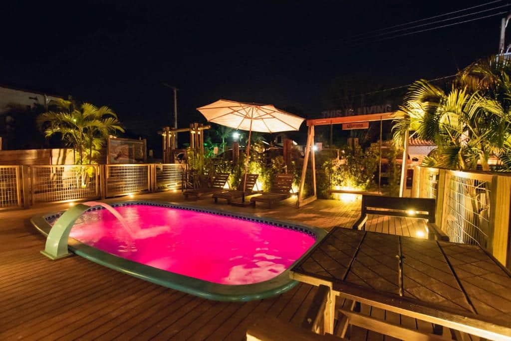 Área externa da Pousada Cacau. Uma piscina no centro iluminada e com cascata, ao redor cadeiras de tomar sol, guarda-sol, um balanço de madeira e luminárias.