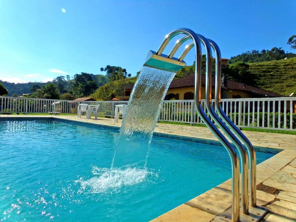 Piscina com água clarinha ao fundo uma casa amarela e algumas árvores durante o dia, ilustrando post pousadas em Visconde de Mauá.