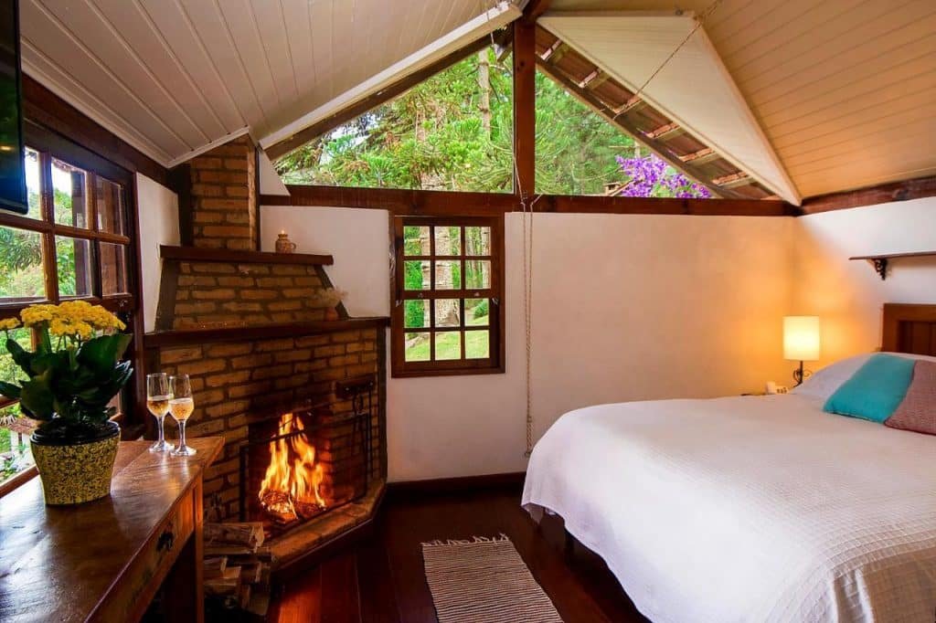 Quarto com cama de casal branca, janelas de vidro com vista para as árvores do lado de fora e lareira dentro do quarto.