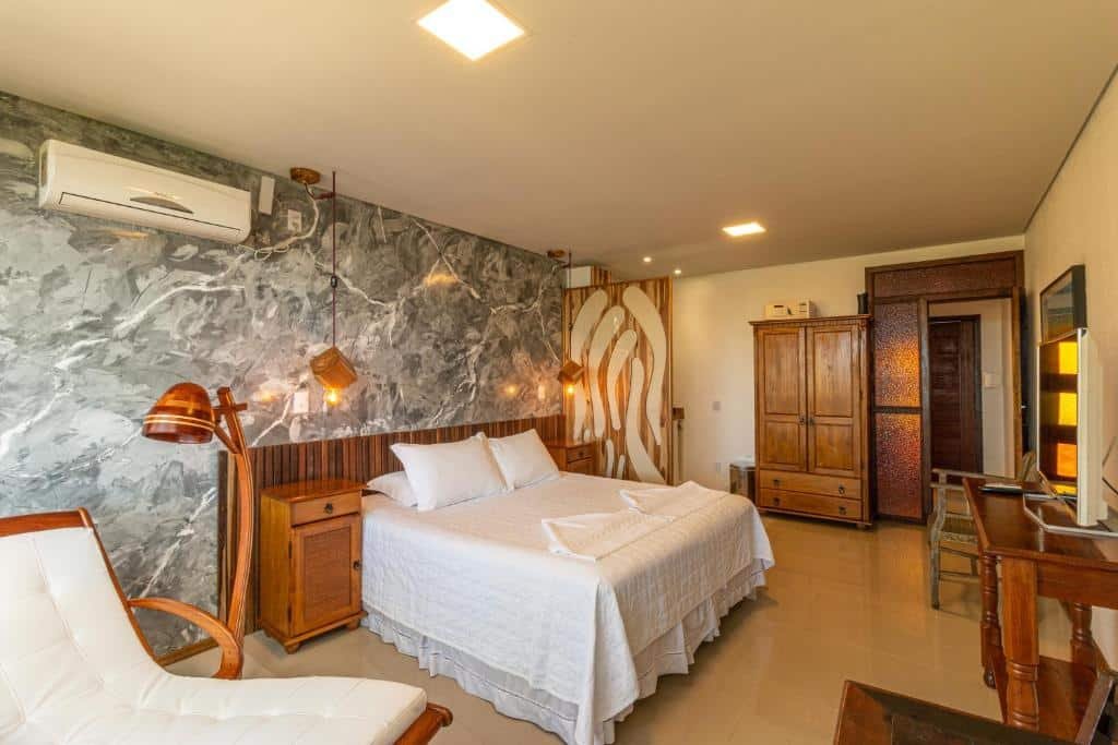 Quarto da Pousada Descanso do Rei. Uma poltrona do lado esquerdo com uma luminária e um ar-condicionado, do lado uma cama de casal com uma cômoda de cada lado. Na direita uma televisão com uma estante e no fundo um guarda-roupa. Foto para ilustrar post sobre pousadas pet friedly em Santa Catarina.