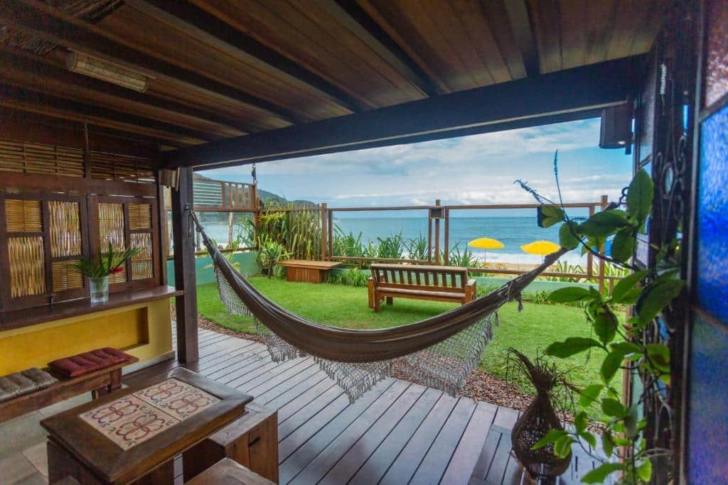 Varanda na Pousada DiMari. Em primeiro plano há uma área coberta em um chão de madeira, com bancos, uma mesinha e uma rede. Ao fundo está um gramado, um banco e uma parede de vidro separando esse espaço da praia.