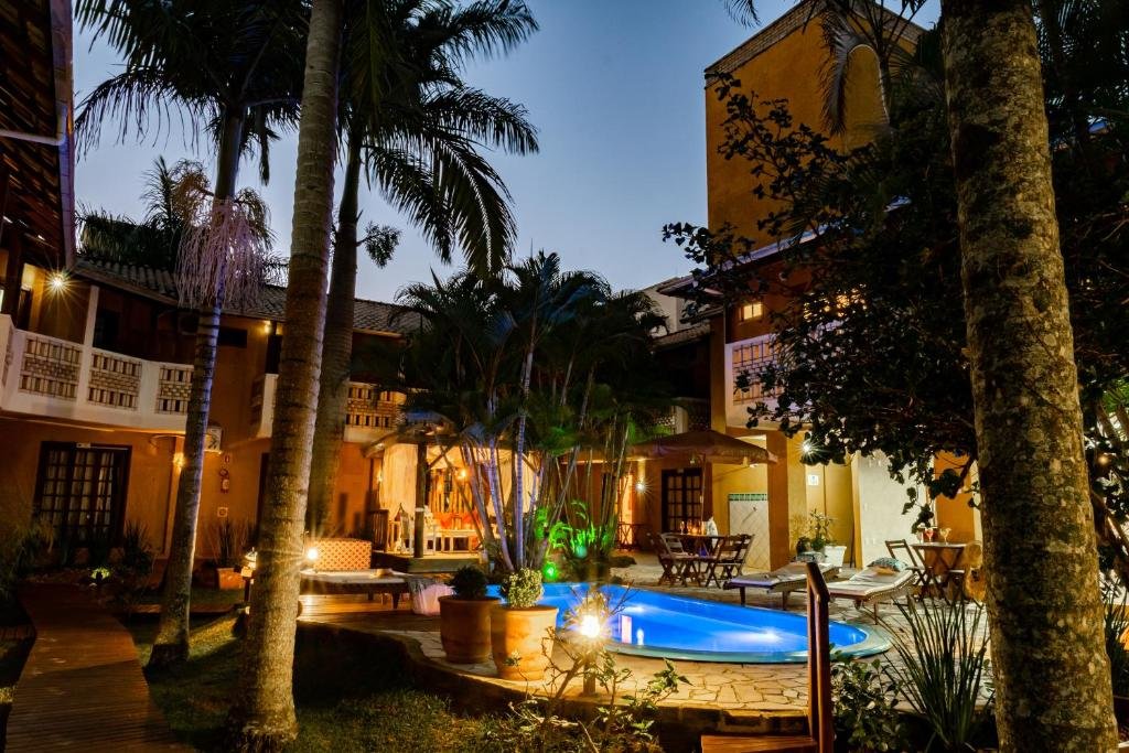 Área externa da Pousada Dom Capudi. Uma piscina no centro iluminada, ao redor cadeiras de tomar sol, jardim e palmeiras.