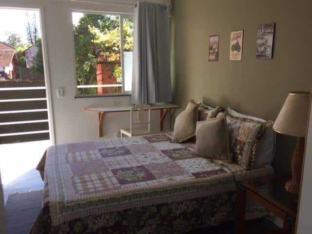 Quarto da Pousada HospedaBia com a cama e parede em tons de marrom, e janela e portas de vidro.
