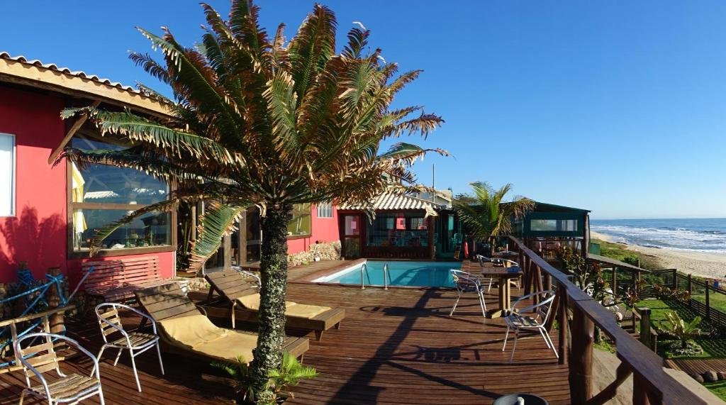 Área externa da Pousada La Luna - Frente Mar. Uma piscina no meio, ao redor cadeiras de tomar sol, cadeiras de aço, mesas de madeira e árvores. Com vista para o mar.