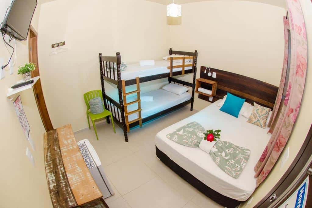 Quarto da Pousada Paraíso Tropical. Do lado direito uma cama de casal, uma cama beliche e uma cadeira. Do lado esquerdo um frigobar, uma estante e uma televisão na parede. Foto para ilustrar post sobre pousadas pet friedly em Santa Catarina.