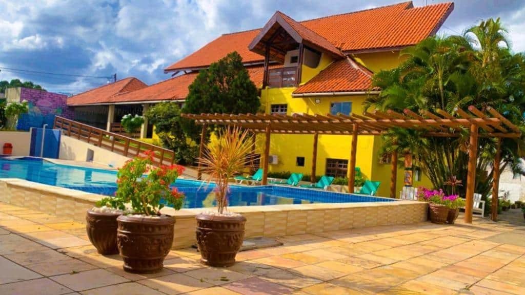 Pousada com paredes amarelas, piscina azul, espreguicadeira e plantas decorativas.