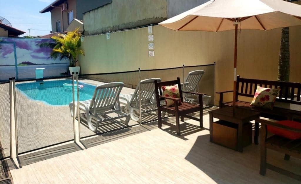 Área externa da Pousada Recanto do Cowboy. Um guarda-sol com bancos de madeira do lado direito, do lado esquerdo uma piscina com cadeiras de tomar sol.