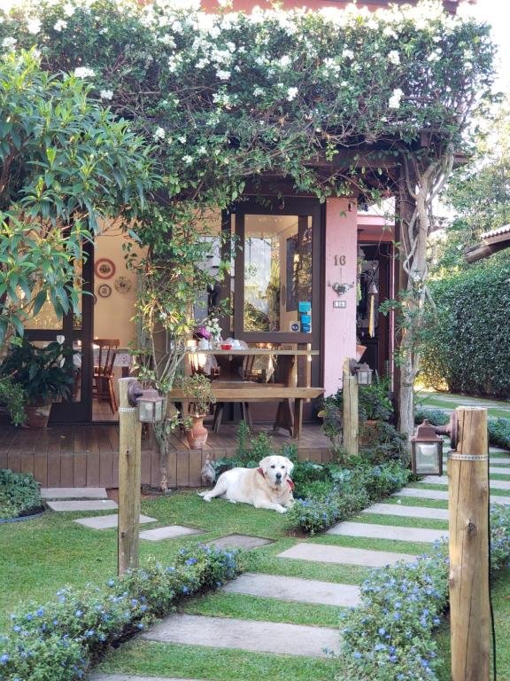 Área externa da Pousada Vila Rosada. Um caminho no jardim, um cachorro sentado, atrás dele o lounge.