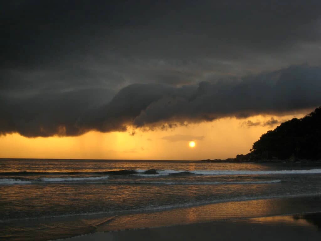 Vista da Praia da Barra do Sahy durante o final do dia com o mar a frente e ao fundo o sol de com várias nuvens em volta.