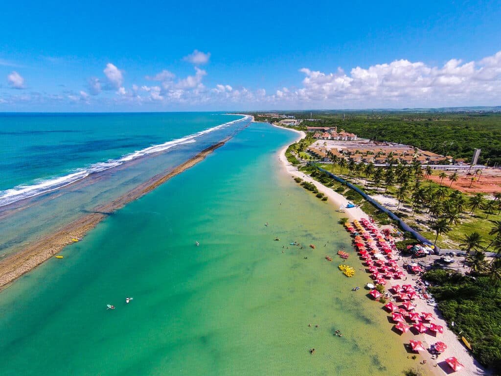 Praia de Muro Alto com uma faixa de areia fina e bem clara, com guarda-sóis vermelhos e brancos, o mar é azul claro com algumas pequenas embarcações