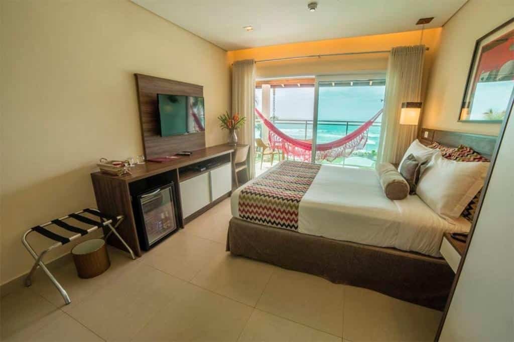 Quarto no Amora Hotel com uma cama de casal, uma varanda dando vista para a praia, com uma rede de descanso presa, do lado de dentro tem uma cama de casal, um móvel com televisão e um frigobar na frente da cama