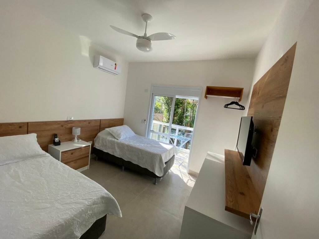 Quarto do Apto Novo Duplex em Juqhey com duas camas de solteiro do lado esquerdo da imagem com uma cômoda no meio delas e em frente as camas uma cômoda com TV presa na parede. Representa airbnb na Barra do Sahy.