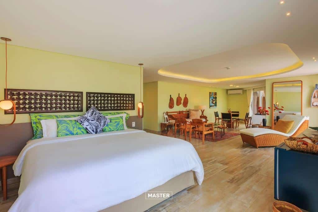 Quarto espaçoso do Armação Resort Porto de Galinhas com uma cama de casal, uma pequena sala de estar com cadeiras e sofás com almofadas, além de uma espreguiçadeira e uma mesa para refeições com quatro lugares