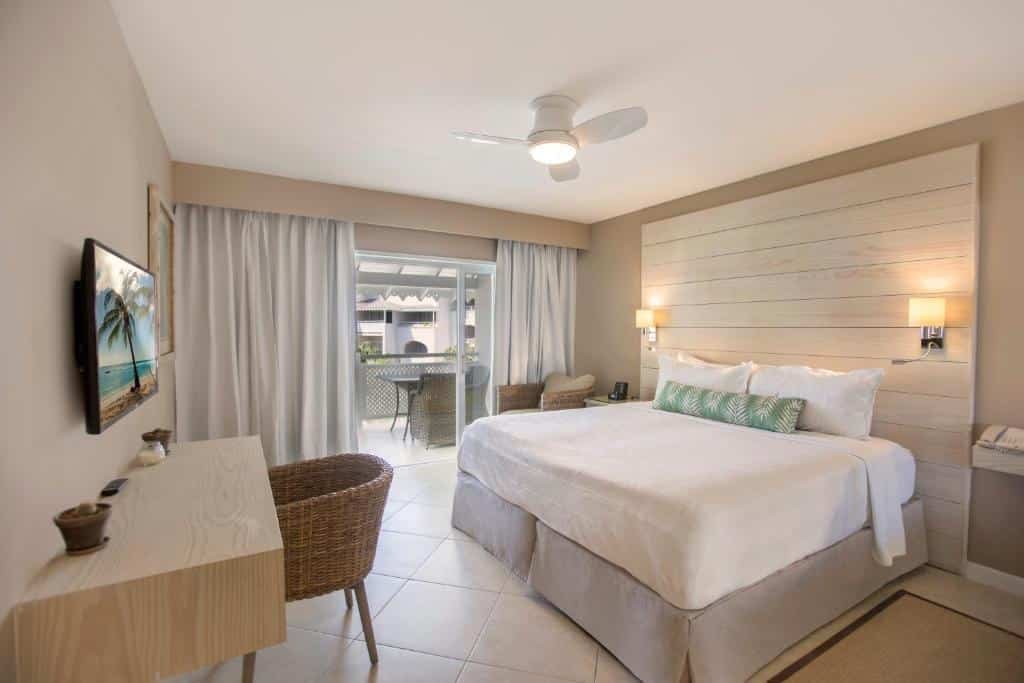 Quarto do Bougainvillea Barbados que mostra uma grande cama de casal e atrás na parede tem um painel de madeira branca com dois abajures. Em frente a cama tem uma mesa de madeira na parede, uma poltrona e uma tv. Já do lado direito do quarto tem uma porta de vidro com cortinas que dá acesso a varanda do quarto.