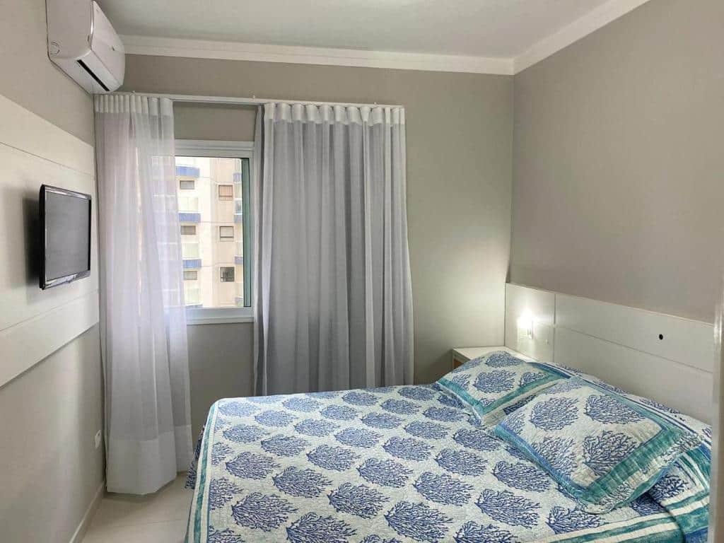 Quarto do Boulevard Riviera Flat, uma cama de casal, uma pequena janela com cortinas e uma televisão, para representar pousadas em Riviera de São Lourenço