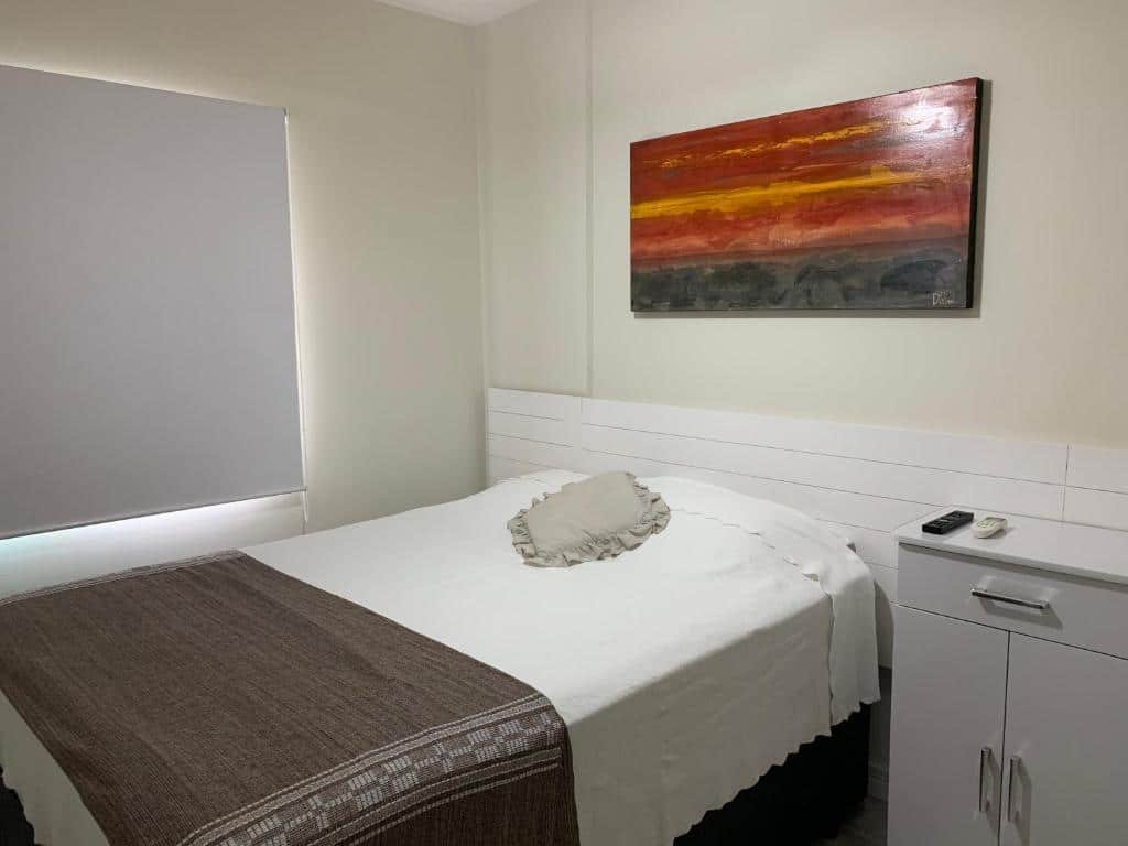 Quarto no Boulevard Riviera Flat - Praia com uma cama de casal, uma pequeno móvel com gavetas, uma janela com persiana e um quadro sob a cama