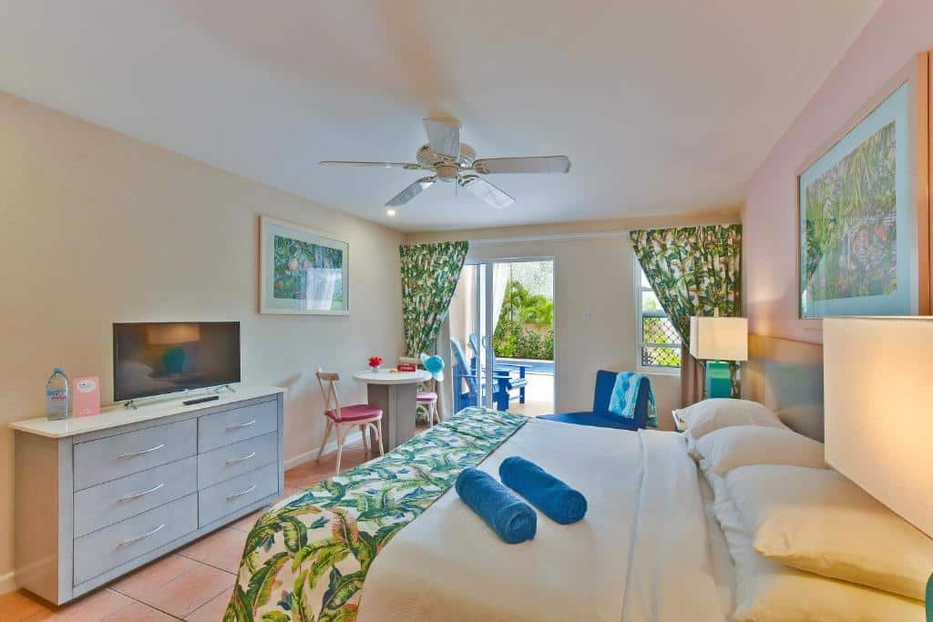 Quarto do Butterfly Beach Hotel. Do lado direito uma cama de casa, de cada lado um abajur e no fundo uma poltrona e uma janela. Do lado esquerdo uma cômoda com uma TV e uma mesa com duas cadeiras. No teto um ventilador.