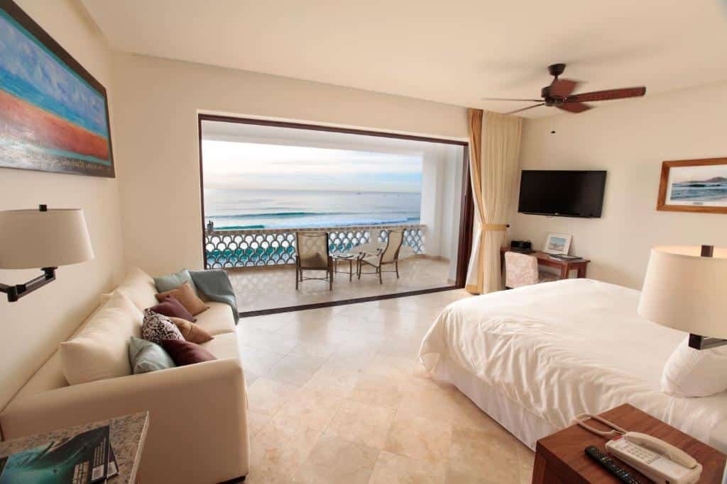 Quarto amplo do Cabo Surf Hotel com cama de casal, sofá do lado direito, TV do lado esquerdo na parede com mesa de trabalho abaixo. E em frente a cama porta ampla de vidro em frente ao mar com varanda com cadeiras.