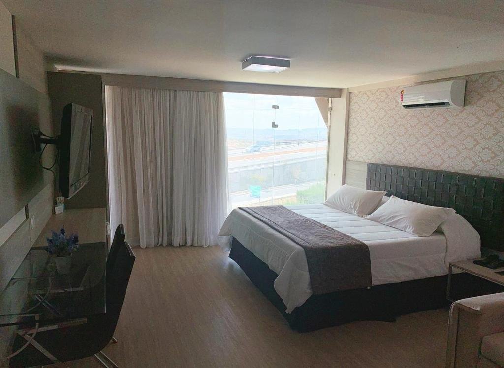 Quarto de hotel com cama de casal com colcha branca e detalhe marrom, cabeceira marrom, ar-condicionado em cima da cama, grande janela ao lado com cortina branca e mesa de vidro em frente a cama com TV em cima. Imagem para ilustrar o post hotéis em Campina Grande.
