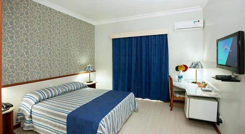 Quarto com cama de casal com colcha azul, papel de parede florido azul, cortina azul, mesa branca, cadeira de madeira, TV e ar-condicionado. Imagem para ilustrar o post hotéis e pousadas em Itu.