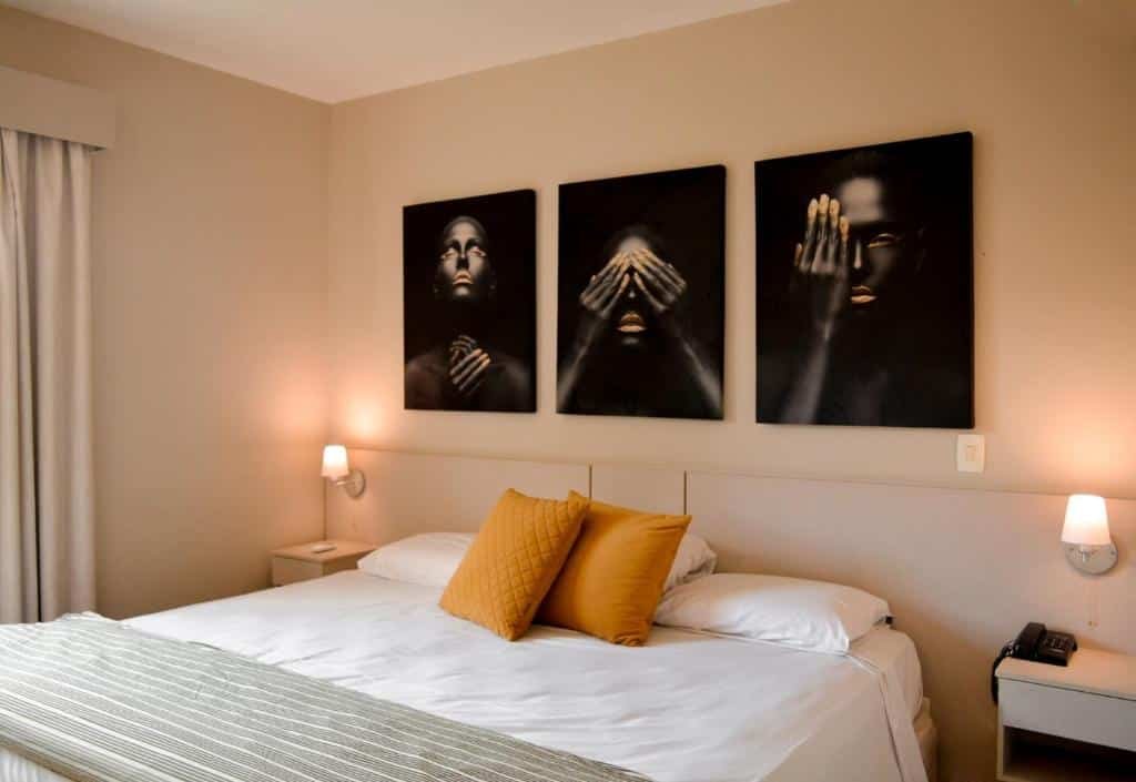 Quarto com cama de casal com colchas brancas, almofada amarela, quadros decorativos em cima da cama e luminárias ao lado da cama. Imagem para ilustrar o post hotéis e pousadas em Itu.
