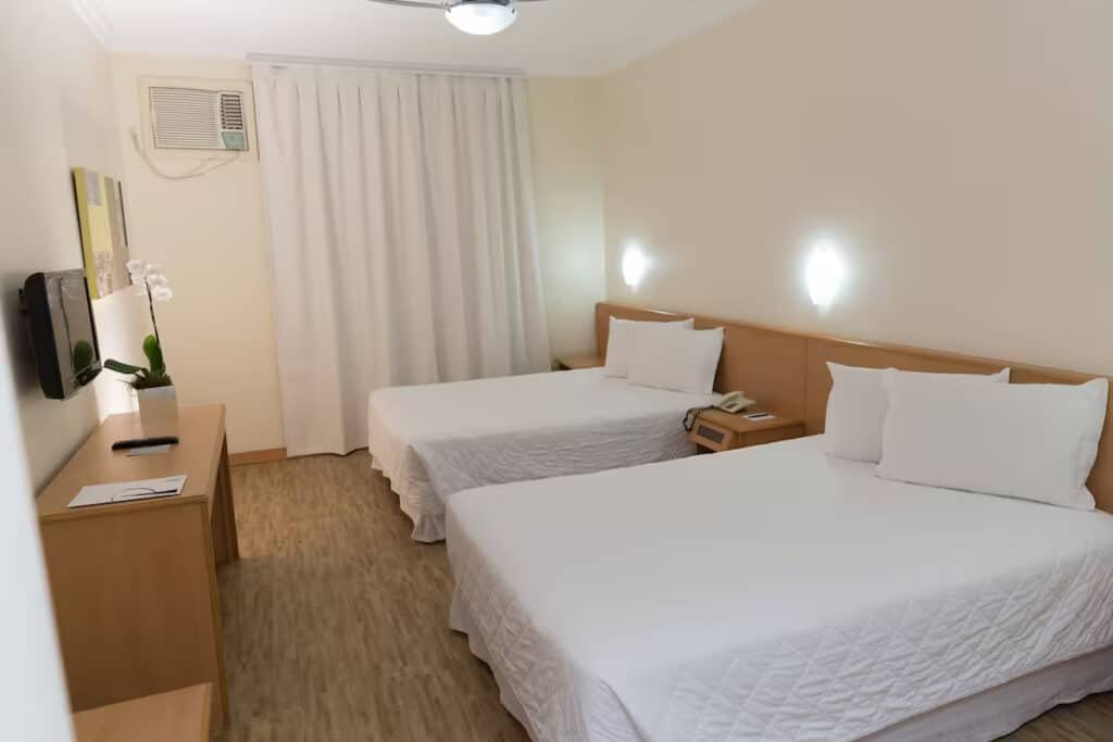 Quarto com paredes e cortinas brancas, duas camas de casal com colcha branca, móvel de madeira, orquídea e TV em frente as camas. Imagem para ilustrar o post hotéis e pousadas em Itu.