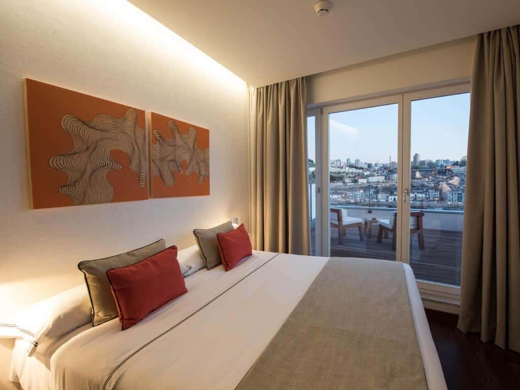 Quarto do Hotel Carris Porto Ribeira com cama de casal do lado esquerdo da imagem, do lado direito da cama portas de vidro que dá acesso a varanda com duas cadeiras com vista para o rio. Representa hotéis com vista do Douro.