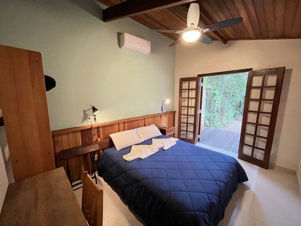 Quarto da Casa Tubarão – A Melhor com mesa de trabalho com cadeira do lado esquerdo da imagem a frente, já no centro do quarto a cama de casal com uma cômoda em cada lado da cama. Representa airbnb na Barra do Sahy.
