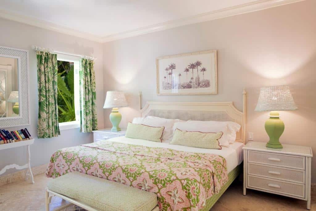 Quarto do Cobblers Cove que mostra uma cama de casal ao centro do quarto, um banco aos pés da cama e de cada lado da cama uma mesa de cabeceira com um abajur. Já do lado direito do quarto tem uma mesa com livros, um espelho e do lado do espelho uma janela aberta com cortinas.
