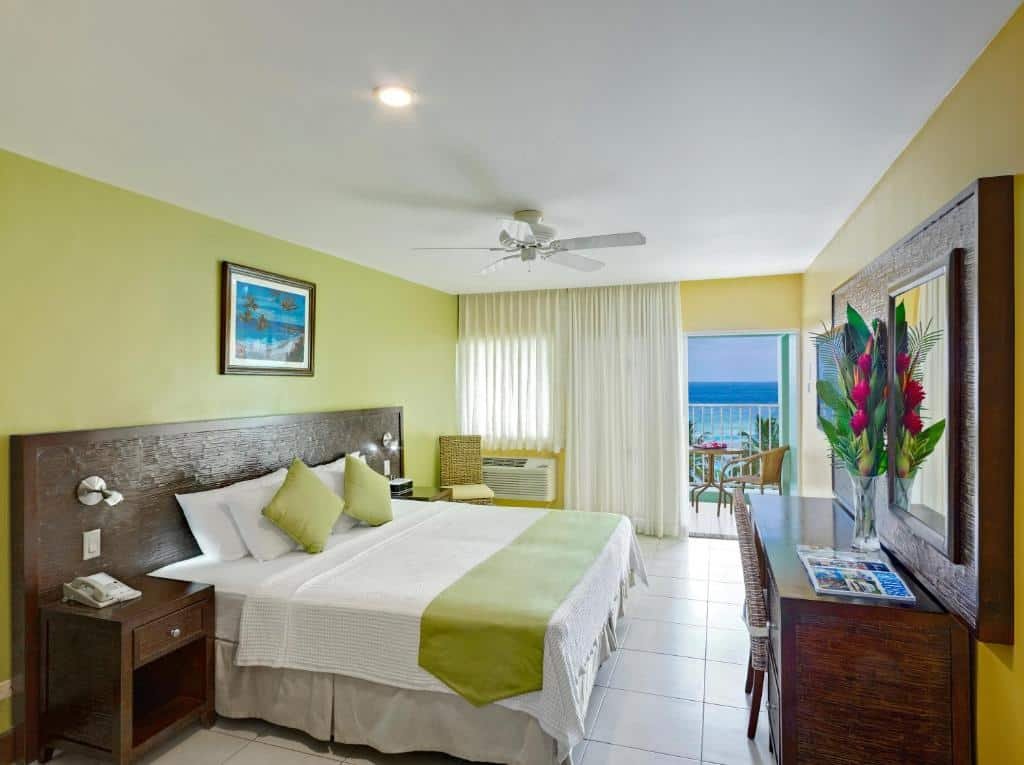 Quarto do Coconut Court Beach Hotel. Uma cama de casal do lado esquerdo, com uma cômoda e um lustre de cada lado. Do lado direito uma mesa de trabalho e uma TV. No fundo a janela com cortina e a porta da varanda. No teto um ventilador.