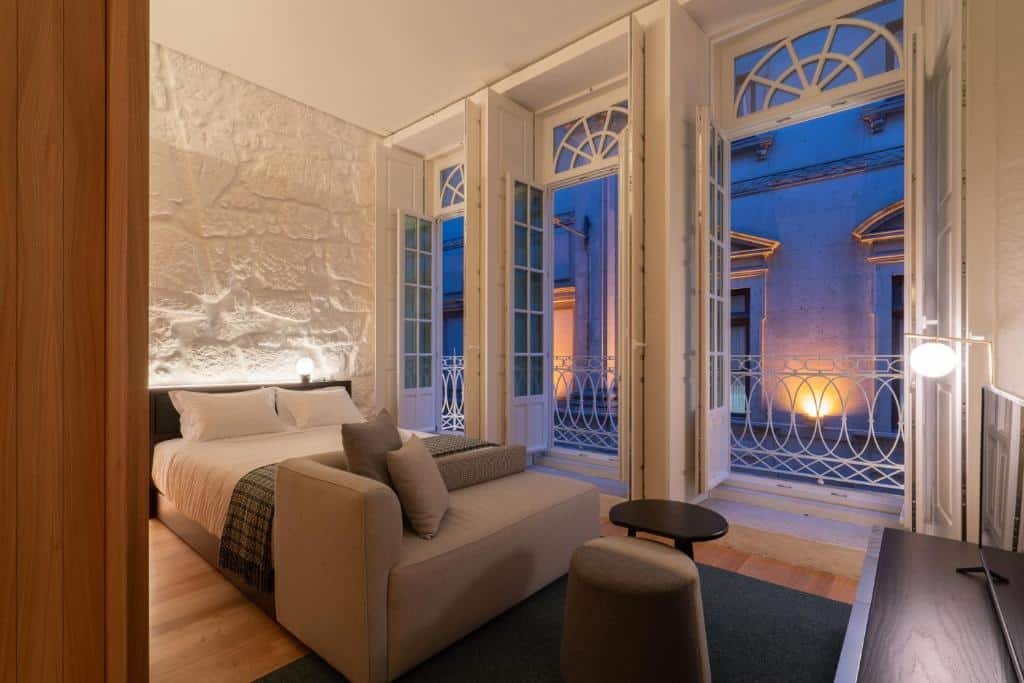 Quarto do d’ALMA Boutique Hotel com cama de casal do lado esquerdo da imagem, com um sofá no pé da cama em frente ao sofá uma cômoda com TV. Representa hotéis no centro do Porto.