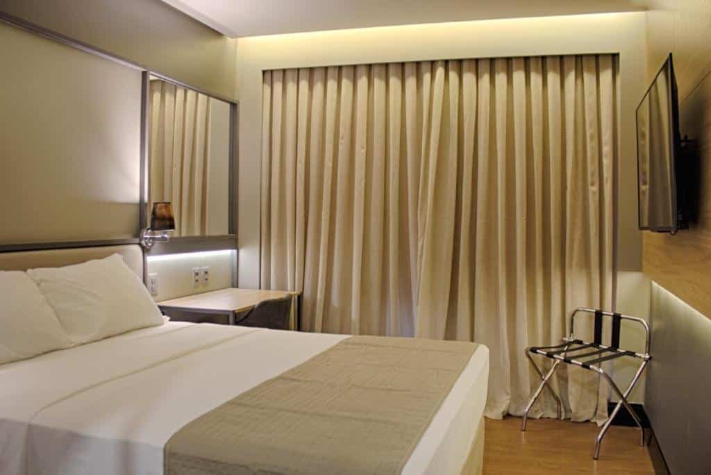 Quarto de hotel com cama de casal com colcha branca e detalhe beje, pequeno espelho e mesa de madeira ao lado da cama, cortina grande beje e TV em frente a cama. Imagem para ilustrar o post hotéis em Campina Grande.