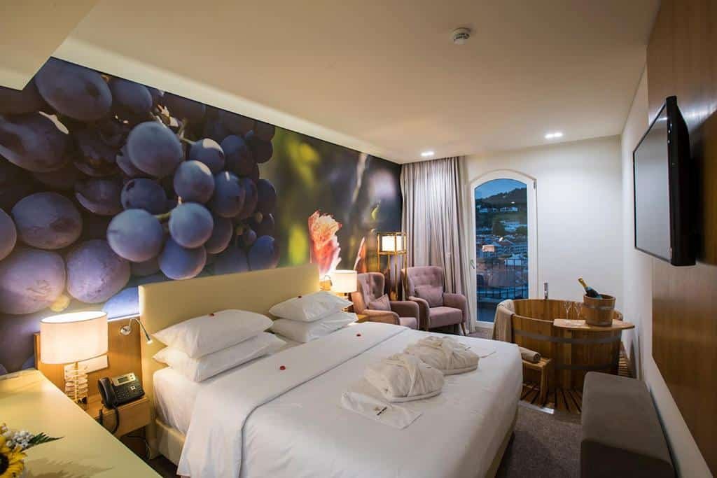 Quarto do Douro Castelo Signature Hotel & Spa com cama de casal do lado esquerdo, duas cômodas ao lado da cama com luminárias, do lado direito da cama duas poltronas e em frente as poltrona uma banheira redonda de madeira de hidromassagem. Representa hotéis no Vale do Douro.