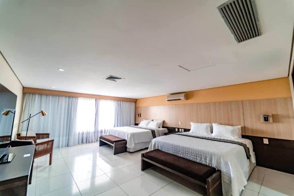 Quarto de hotel bem amplo com duas camas de casal, cadeiras de madeira, mesa com TV, grande janela com cortinas brancas e cabeceiras de madeira em cima das camas. Imagem para ilustrar o post hotéis em Campina Grande.
