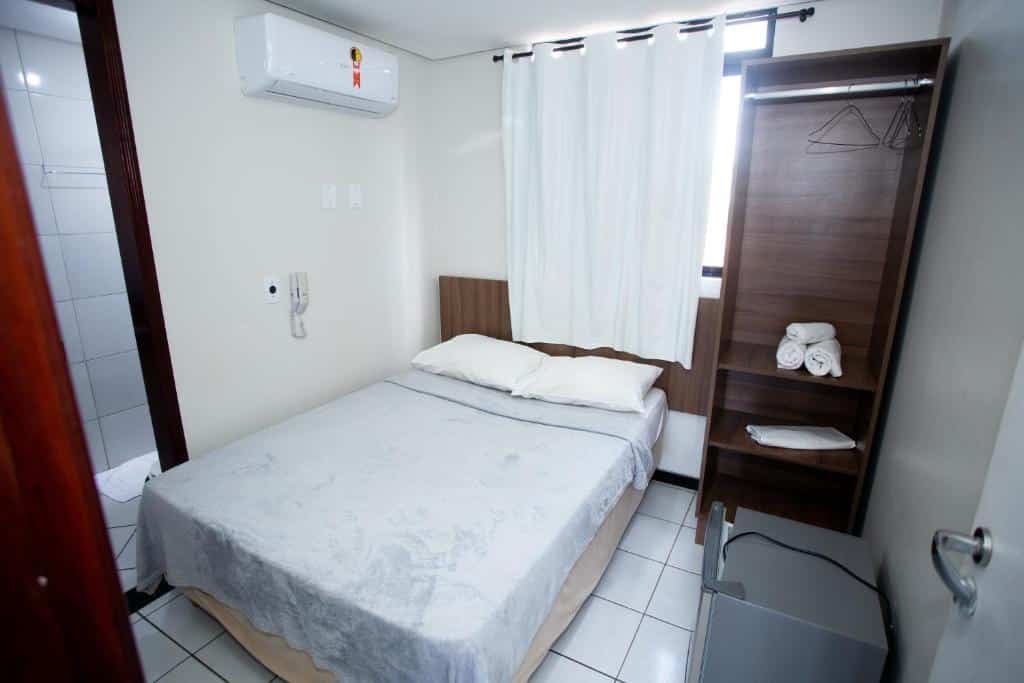 Quarto de hotel simples com cama de casal, paredes brancas, ar-condicionado, cortina branca, cabeceira de madeira e pequeno armário de madeira com toalhas e porta para o banheiro. Imagem para ilustrar o post hotéis em Campina Grande.