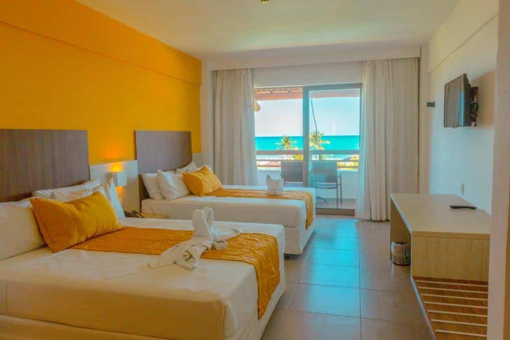 Quarto do Enotel Porto de Galinhas com duas camas, uma de casal e um de solteiro, com uma varanda dando vista para a praia, e há uma televisão presa na parede de frente para as camas