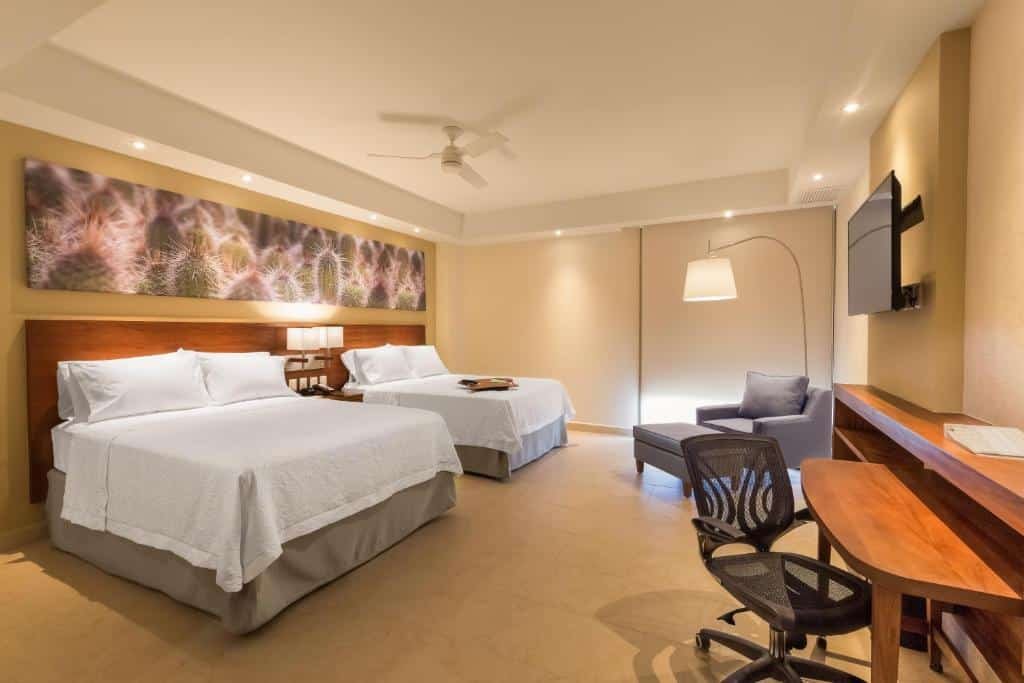 Quarto do Hampton Inn & Suites by Hilton Los Cabos com duas camas de casal e uma cômoda no meio com luminária. Poltrona cinza ao lado esquerdo. TV em frente as camas presa na parede com mesa de trabalho e cadeira abaixo.