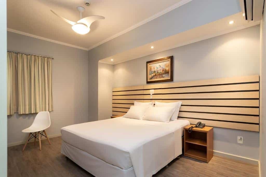 Quarto com cama de casal com lençol branco centralizada, cabeceira marrom clara, quadro decorativo acima da cabeceira, cadeira branca ao lado da cama e cortinas bege. Imagem para ilustrar o post hotéis e pousadas em Itu.