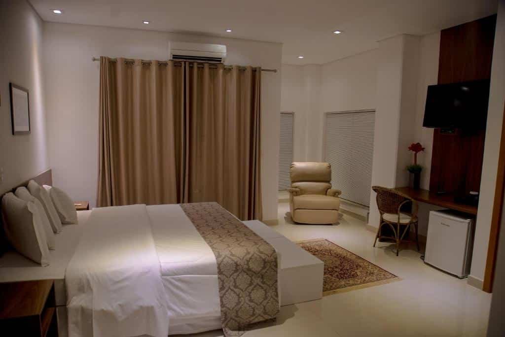 Quarto luxuoso com cama de casal com colcha branca, mesa, cadeira, frigobar e TV em frente a cama, poltrona bege, cortina marrom claro e ar-condicionado. Imagem para ilustrar o post hotéis e pousadas em Itu.