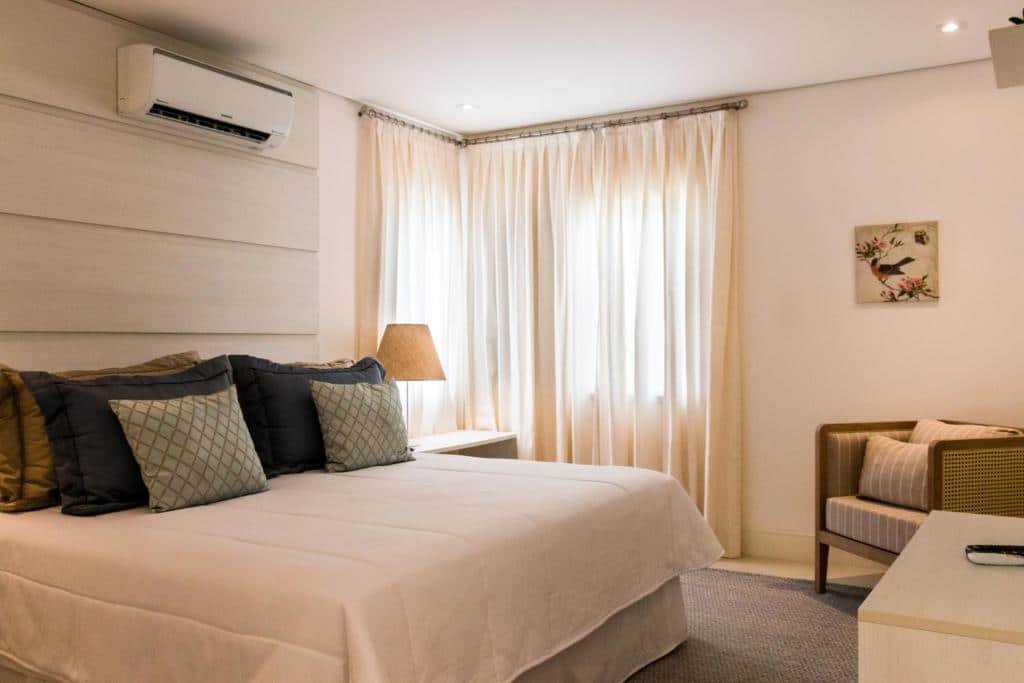 Quarto do Hotel Maui Maresias com uma cama de casal, uma janela com cortinas, uma poltrona, um tapete e muitas almofadas sob a cama