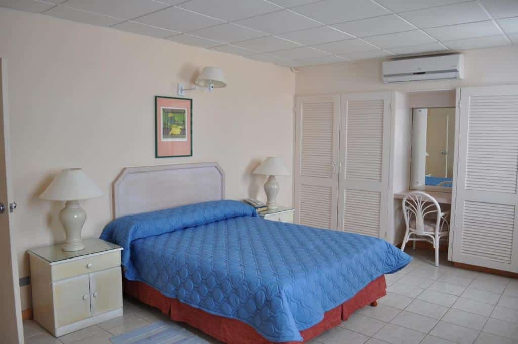 Quarto do Hotel PomMarine. Uma cama de casal, de cada lado uma cômoda e um abajur. No fundo, o armários, no centro uma mesa de trabalho, espelho e ar-condicionado.