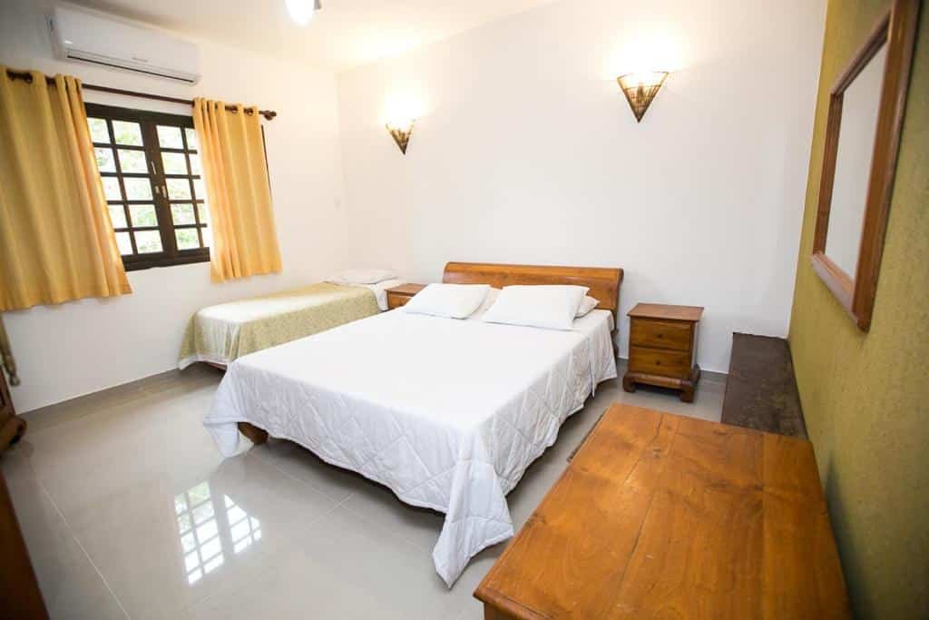 Quarto da Hotel Pousada dos Condes com uma cama de casal e uma de solteiro, com uma janela com cortinas, um espelho, uma cômoda e um ar-condicionado de teto