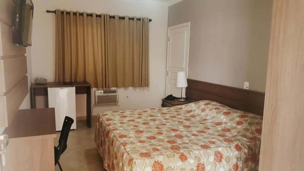 Quarto com cama de casal com colcha florida, mesa de madeira e TV em frente a cama, frigobar, ar-condicionado e cortina marrom ao lado da cama. Imagem para ilustrar o post hotéis e pousadas em Itu.