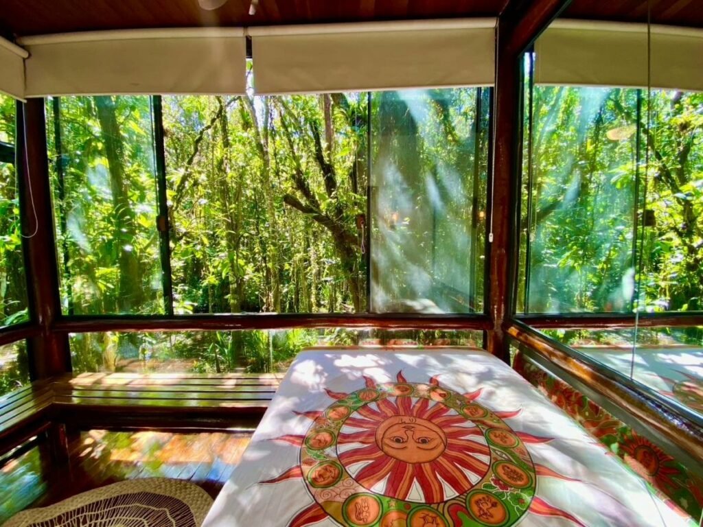 Quarto do Housing 31 durante o dia com cama de casal do lado direito da imagem as paredes são de vidro que tem vista para a natureza. Representa airbnb em São Sebastião.