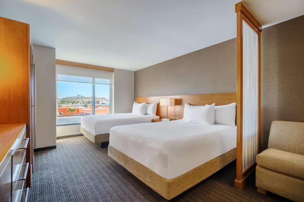 Quarto do Hyatt Place Los Cabos com duas camas de solteiro, janela ampla e poltrona.